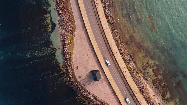 Aerial view of a curved coastal road and sea. High angle shot of a road with white cars next to a traditional windmill and rocky shoreline.