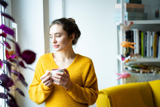 Woman in yellow sweater holding a cup and looking out the window in a cozy home setting