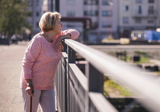 Smiling senior woman with walking stick outdoors leaning on handrail in city