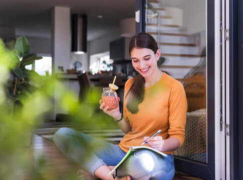 Smiling young woman writing in notebook with drink at home by open door
