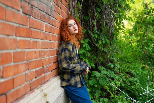 Woman with red curly hair in jeans leaning on brick wall outdoors