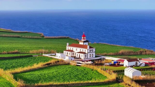 Aerial view of Farol da Ponta do Arnel lighthouse perched on a lush green cliff above the deep blue Atlantic Ocean in the Azores, Portugal.