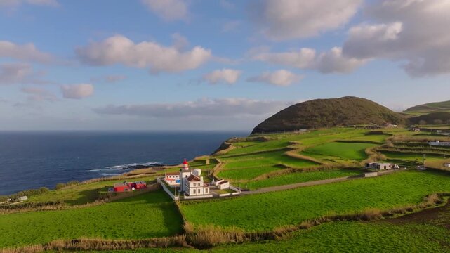 Aerial view of Farol da Ponta do Arnel lighthouse perched on a lush green cliff above the deep blue Atlantic Ocean in the Azores, Portugal.