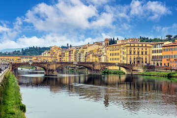 Fototapeta premium The city of Florence with the Arno River flowing beneath the Santa Trinita Bridge. Buildings line the riverbank, and the sky is bright with clouds.