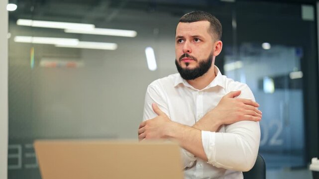 Focused businessman experiences cold while working on laptop in modern office. Feeling discomfort, adjusts air conditioning temperature using remote control for improved climate comfort.