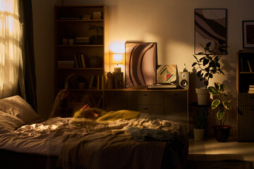 Young woman lying on bed in softly lit bedroom, relaxing with hands behind head, surrounded by bookshelves, plants, framed artwork, and audio speakers © Mediaphotos