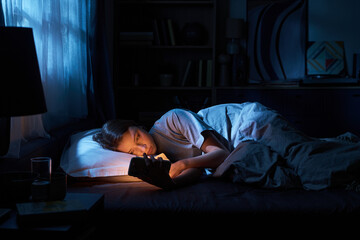 Portrait of teenage girl lying in bed at night holding smartphone close to face, illuminated by screen light, gazing at device with tired expression, bedroom background © Mediaphotos