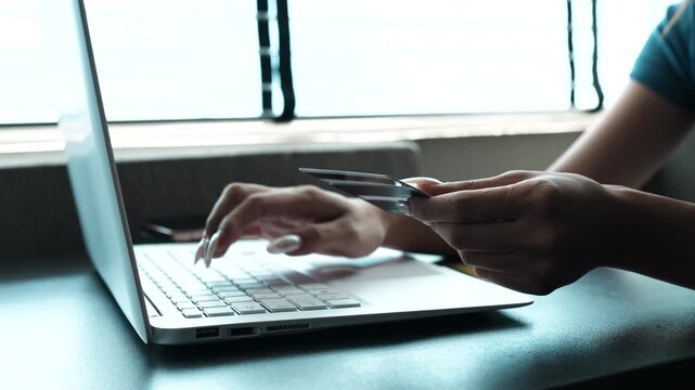 A woman holding a credit card while using a smartphone for online shopping, sitting at a desk with a laptop, representing digital payments and modern e-commerce lifestyle.