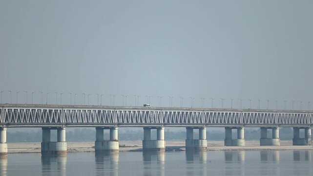 A steel truss bridge spans across a calm river, supported by concrete pillars with soft reflections in the water. The clear sky and open landscape create a peaceful and minimal scenic view.