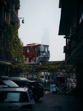 View of a red and white train gliding above the street, framed by buildings and trailing vines, under a misty sky, Chongqing, Chongqing, China.