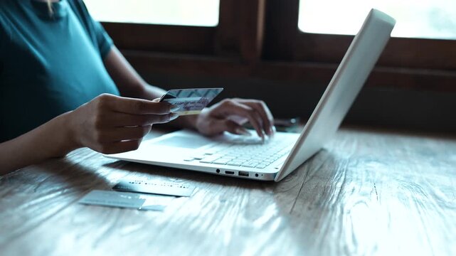 A woman holding a credit card while using a smartphone for online shopping, sitting at a desk with a laptop, representing digital payments and modern e-commerce lifestyle.