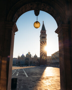 View of a sunburst behind the Campanile di San Marco, framed by an archway in the Piazza San Marco, casting shadows across the square, Venice, Veneto, Italy.