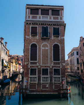 View of a tall, narrow brick building rising beside a canal, its arched windows reflecting the serene water, a timeless scene, Venice, Veneto, Italy.