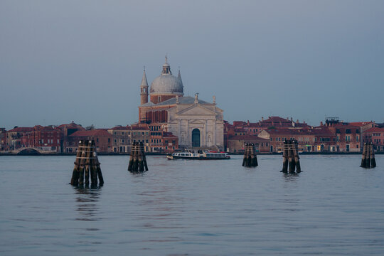 View of weathered wooden posts rise from the tranquil waters, framing the distant, ornate architecture under a soft sky, Venice, Veneto, Italy.