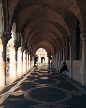 View of the arched corridor, where light and shadow dance on the patterned floor, leading to a distant square, , Venice, Veneto, Italy.