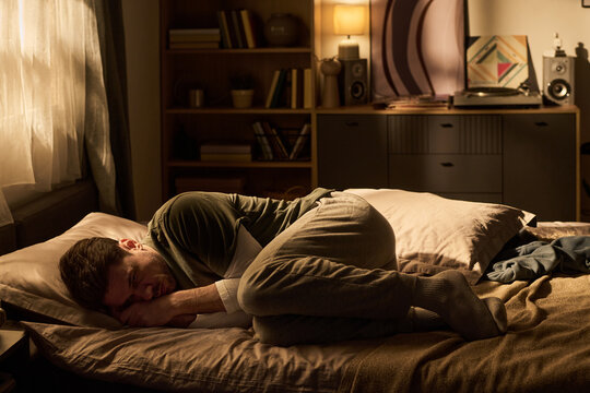 Young man lying curled up on bed facing window, resting head on pillow with eyes closed, appearing to be sleeping or napping in softly lit bedroom interior