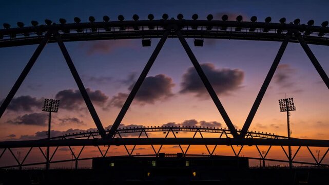 Framing curved steel roof truss and seating bowl, sunset deepening, clouds drifting, towers glowing