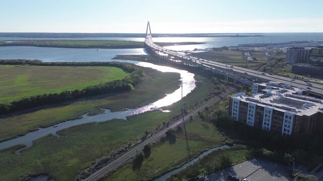 Cooper River marshlands sit below Arthur Ravenel Jr. Bridge beside Morrison Dr and NoMo corridor. Bright concrete, crisp shadows, textured wetlands contrast with dense rooftops in South Carolina