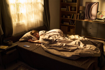 Young woman sleeping in bed with hand resting on pillow, smartphone lying on sheet near her, morning sunlight streaming through window, bookshelf in background © Mediaphotos