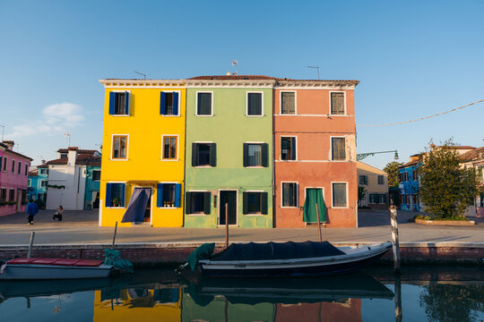 View of vibrant yellow, green, and peach houses reflecting in the canal's still waters, framed by boats and a serene sky, Burano, Veneto, Italy.