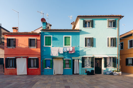 View of vibrantly painted houses in a row, each a different hue of orange, blue, and green, beneath a clear sky, Burano, Veneto, Italy.