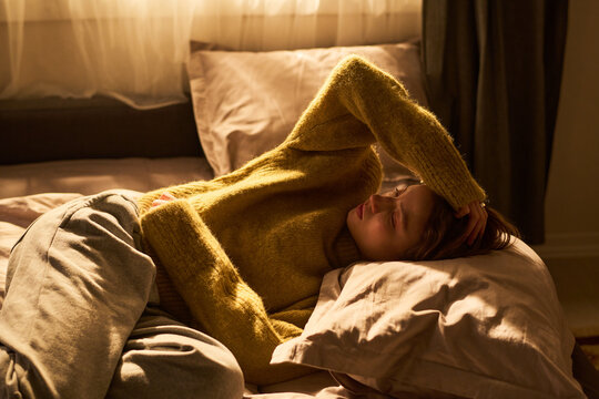 Young woman lying on bed holding head with hand, appearing to experience discomfort or pain, resting on pillow in softly lit bedroom during daytime
