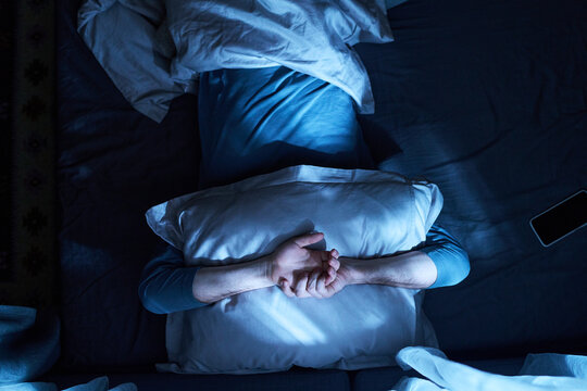 Adult man lying in bed covering face with pillow, crossing arms over pillow, smartphone resting nearby on bed, appearing restless or unable to sleep at night