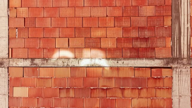 Workers are building a wall using red bricks. The bricks are arranged in horizontal rows, showing a clear pattern. Dust and debris surround the area, suggesting ongoing construction.