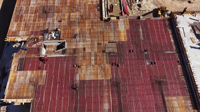 Workers are busy on a construction site, placing steel bars to prepare for a concrete pour. The area shows a large framework and materials scattered around as work continues.