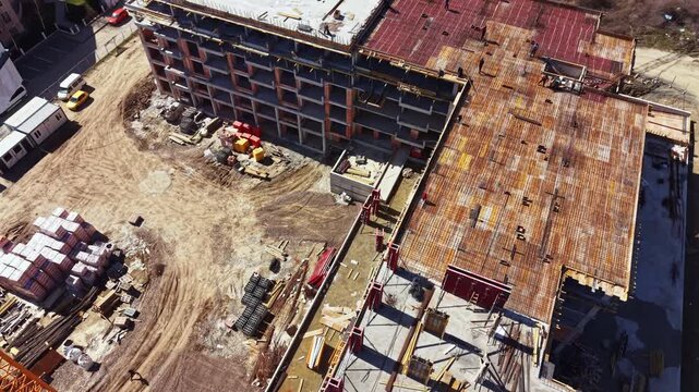Workers and machinery are active at a construction site. Two large buildings are in various stages of completion. Materials are scattered around the area in a busy environment.