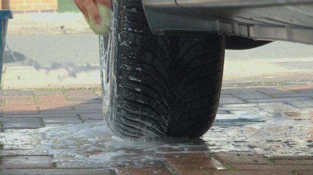 Unrecognizable man cleaning car wheel with sponge at home driveway, saving money through DIY vehicle washing. Close up of tire scrubbing with soap foam on pavement, domestic car care routine and