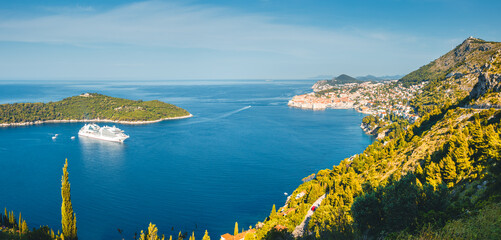 Obraz premium Cruise ship sailing into the stunning turquoise waters of Dubrovnik, with lush hills and historic townscape on a sunny day. Croatia, Dalmatia, Europe.