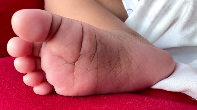 Close-up video of a newborn baby&rsquo;s soft foot resting on a red fabric surface, showing delicate skin texture and tiny toes. Warm natural lighting highlights innocence, comfort, and early childhood det