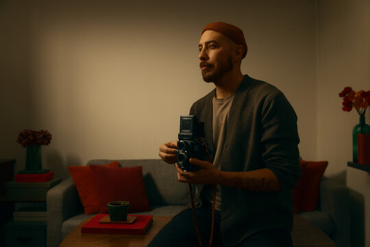 A man in a beanie and cardigan sits in a warm, cozy room holding a vintage twin-lens reflex camera. This moody, cinematic portrait captures a nostalgic, creative atmosphere with soft indoor lighting.