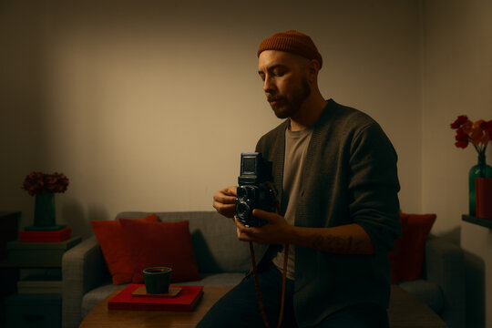 A bearded man in a warm beanie and cardigan carefully handles a vintage twin-lens reflex camera in a cozy, moody living room with soft lighting, red accents, and a mid-century modern aesthetic.
