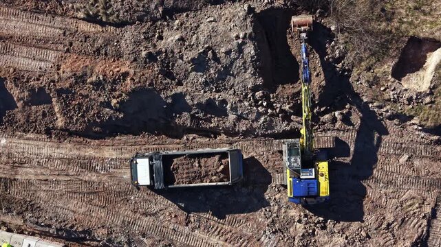 Heavy machinery works at a construction site to move dirt and prepare the ground. A truck is filled with soil while an excavator digs nearby during the day.