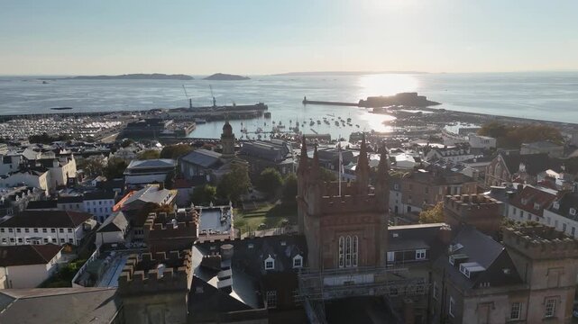 St Peter Port Guernsey sunset flight over Elizabeth College , St James and town rooftops towards the harbour with sun setting over Castle Cornet, Herm and Jethou on calm Summer evening