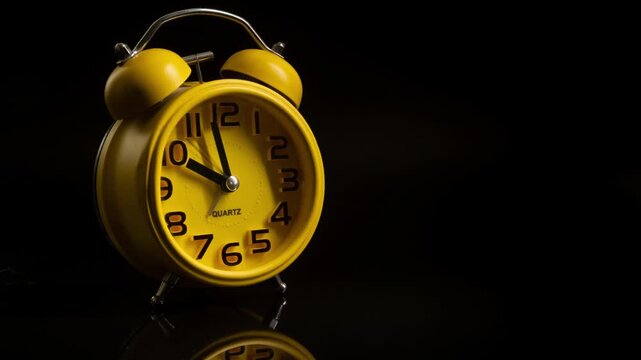 Close up timelapse video of a yellow bedside alarm clock showing the hands moving rapidly against a black background.