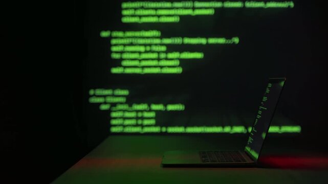 Laptop in dark room with glowing green programming code projected behind screen. Hacking, cybersecurity threats, cybercrime, and digital intrusion technology concept. Space for text