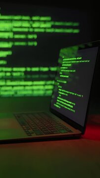 Lines of programming code run on screen of laptop standing on table in dark room, with green glowing hacker script projected in the background. Hacking, cyber attack and cybersecurity threat concept.