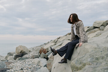 Young woman sits on rocky shore wearing casual clothes, looking away and smiling. Overcast sky...
