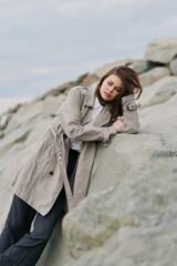 Young woman in knitted coat leans on rocks outdoors, eyes closed, enjoying peaceful moment. Calm...