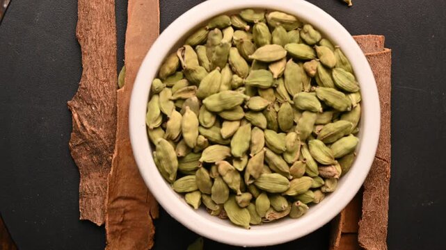 Top View of Cardamom (Elechi) in White Bowl with Cinnamon Sticks (Dalchini) on Black Background &ndash; Rotating Footage