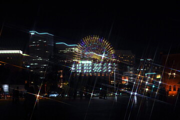 夜景に輝く観覧車と高層ビル群、そして光の軌跡。都会の夜の賑わいと華やかな雰囲気を捉えた一枚。 © BRUCE