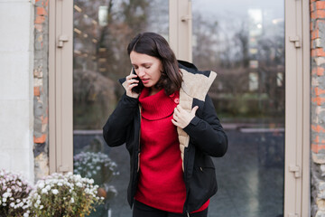 Fototapeta premium Caucasian woman in her 40s with dark hair wearing warm clothes talking on the phone outdoors during her working time. Mature female managing her routine or personal tasks and breathing fresh air.