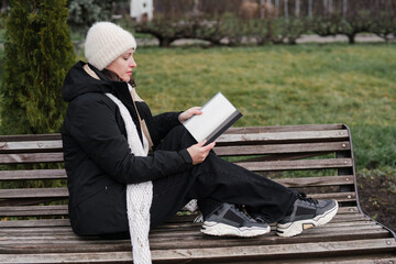 Fototapeta premium Caucasian woman in her 40s with dark hair wearing warm clothes reading a book outdoors in autumn park. Mature female enjoying her personal time and leisure activity, relaxing and breathing fresh air.