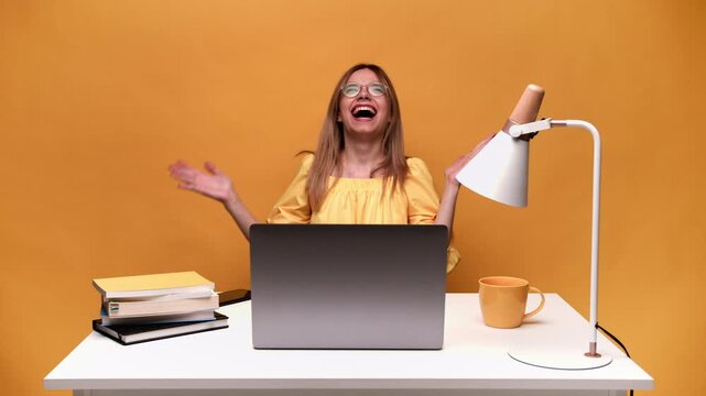 Overjoyed young woman laughing hysterically while working on her laptop against a vibrant yellow background in a studio setting