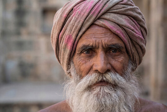Close-up portrait of an elderly Indian man with a long white beard and traditional turban looking directly at the camera against a blurred stone background.