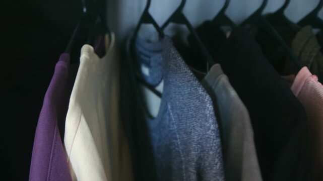 Man hand selecting outfit from rail t shirts hanging in dark closet, highlighting daily routine of choosing what wear from casual clothing collection. Person choosing clothes from wardrobe hanger rail