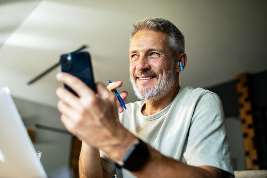 Smiling mature man on smartphone with earbuds at home office